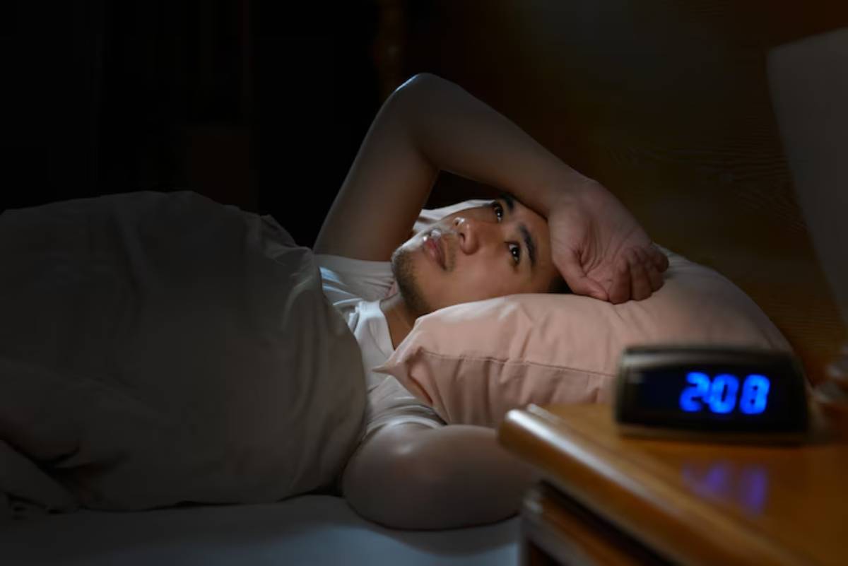 A person lying in bed under a blanket, with a pink pillow nearby and a digital clock displaying 2:08 AM on the bedside table.
