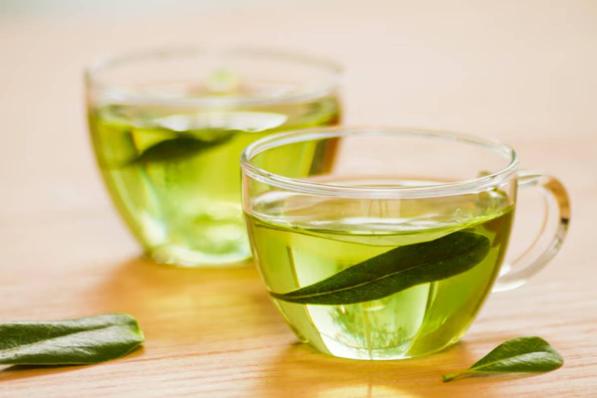 Two clear cups of vibrant green tea, each garnished with a leaf, sit on a wooden surface. Natural light enhances their freshness.