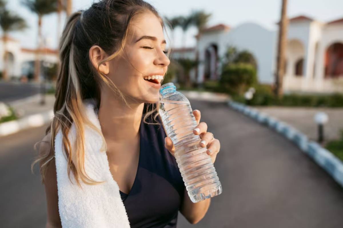 A person holds a water bottle and a towel around their neck, walking on a sunny street lined with palm trees and buildings.