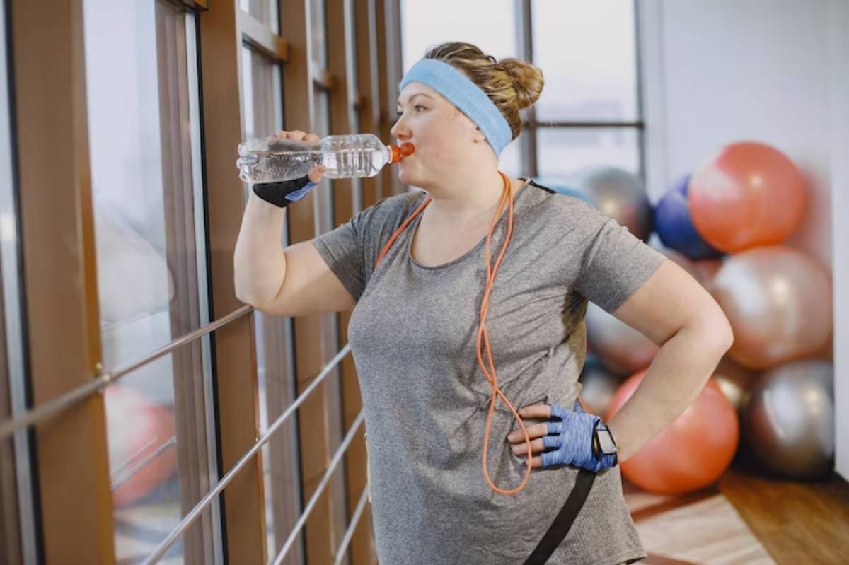 A fit person in a gym, wearing a gray t-shirt and blue accessories, takes a break to sip water from a bottle. Exercise equipment is visible in the background.