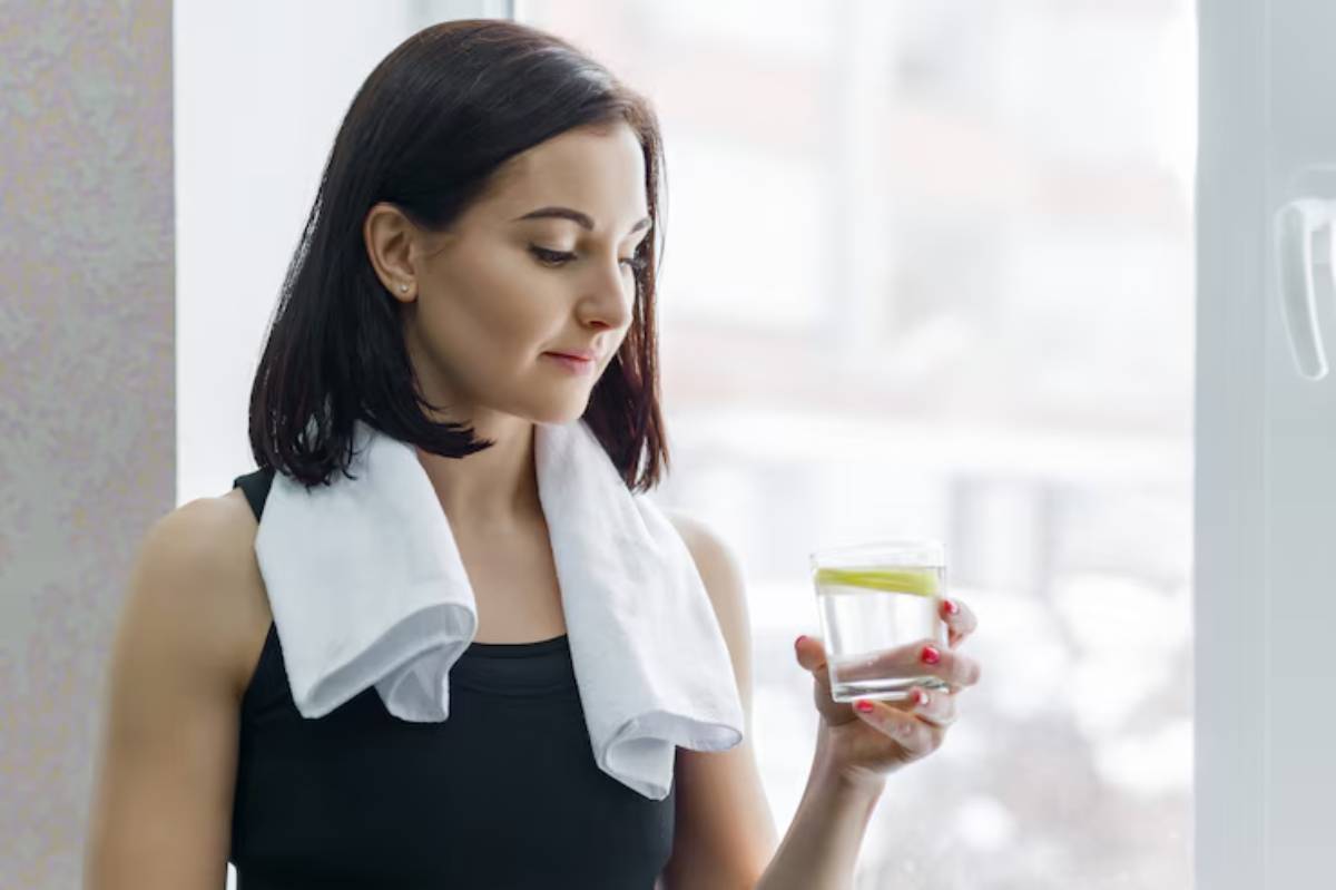 A person holding a glass of water with a lemon slice, wearing a black tank top and a towel around their neck, stands by a window.