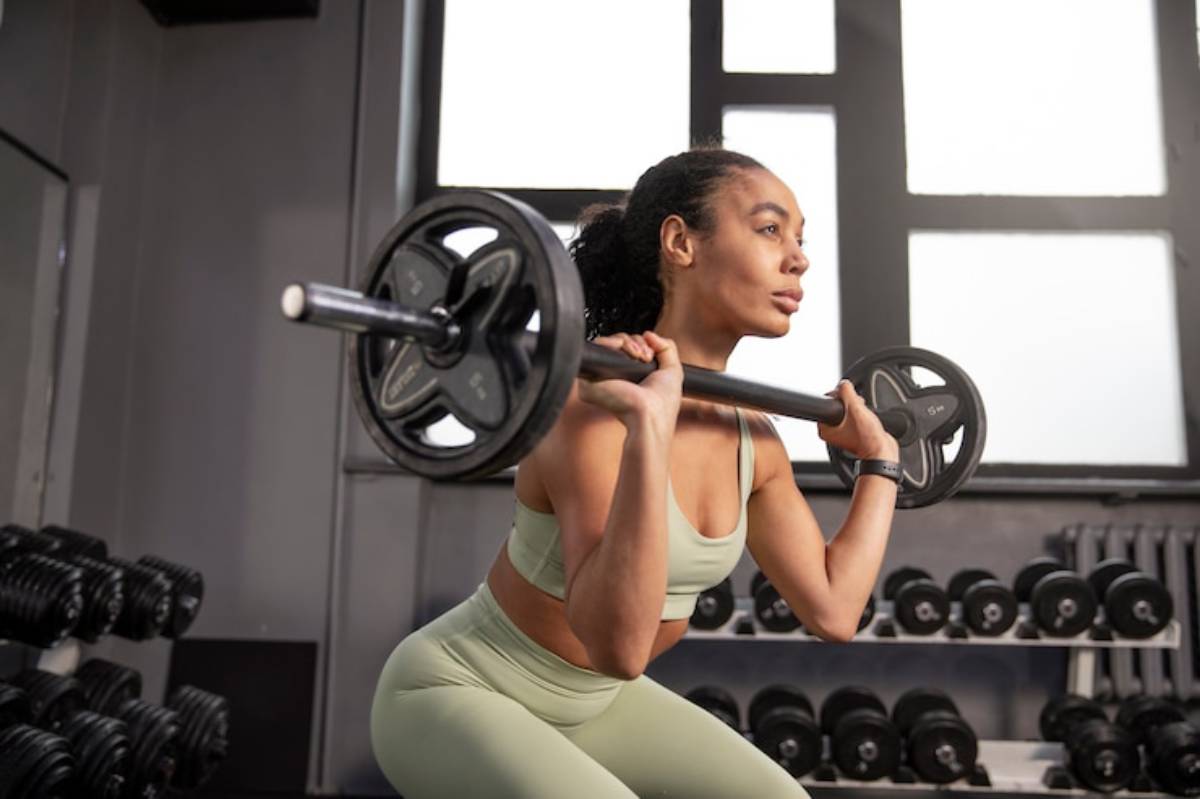 A woman in activewear performs a squat with a barbell in a gym, surrounded by weights and large windows letting in natural light.