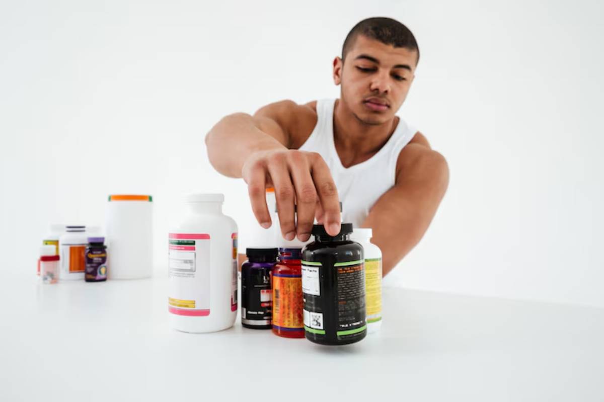 A person in a white tank top reaches for various bottles of dietary supplements on a clean, white surface.