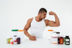 A muscular person in a white tank top poses with a strong arm, surrounded by various bottles of dietary supplements on a white table.