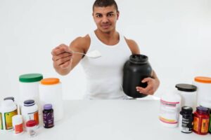 A person holding a black container and a spoonful of powder, surrounded by various fitness supplements on a white background.