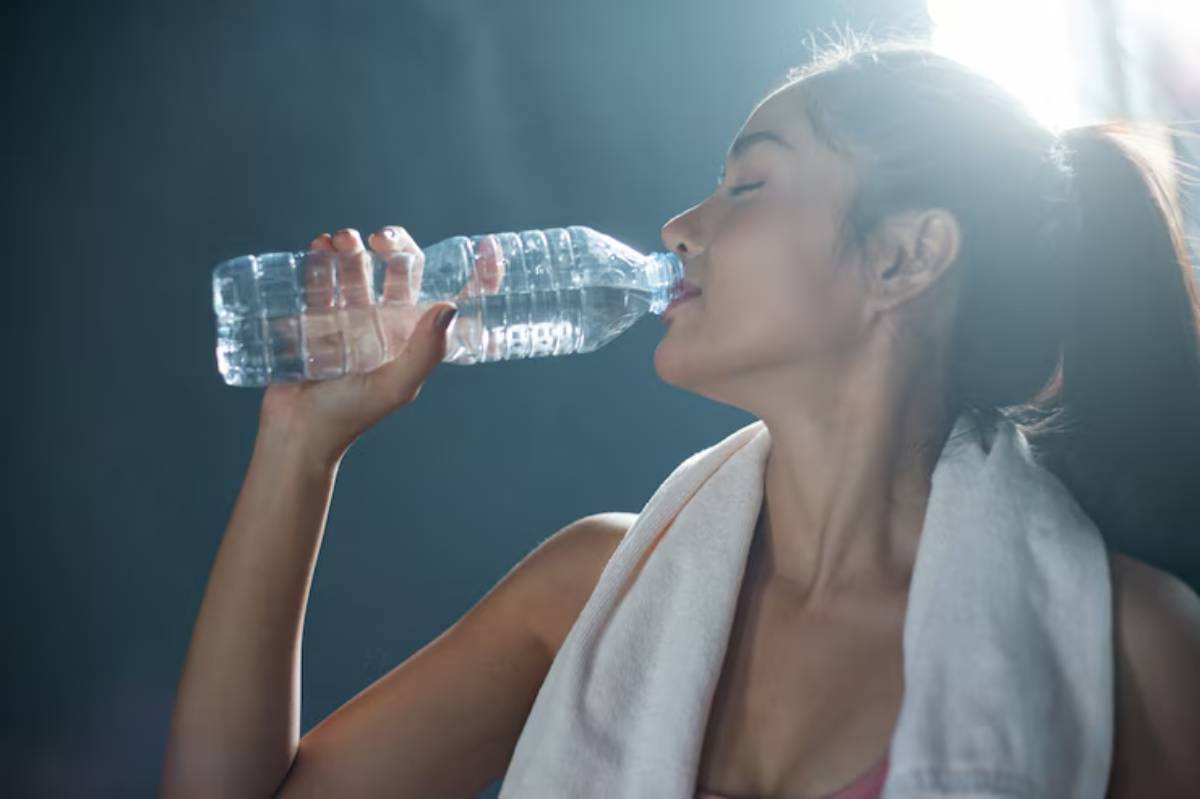 A person drinks from a plastic water bottle, glistening with light, wearing a towel around their neck, in a moody, dimly lit environment.