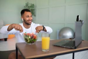 A person sitting at a table, enjoying a salad and orange juice, with a laptop and gym equipment in a light, modern room.