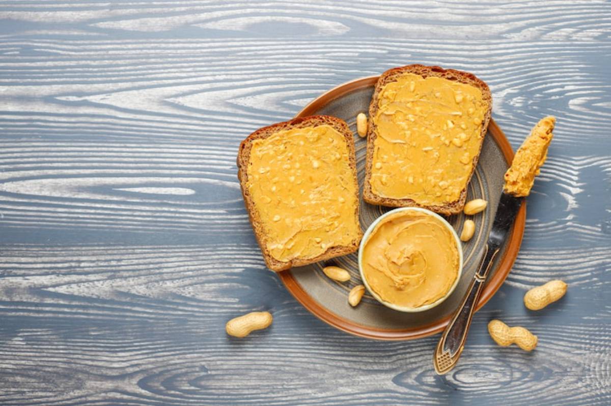 Two slices of bread spread with peanut butter on a blue wooden surface, accompanied by a bowl of peanut butter and scattered peanuts.