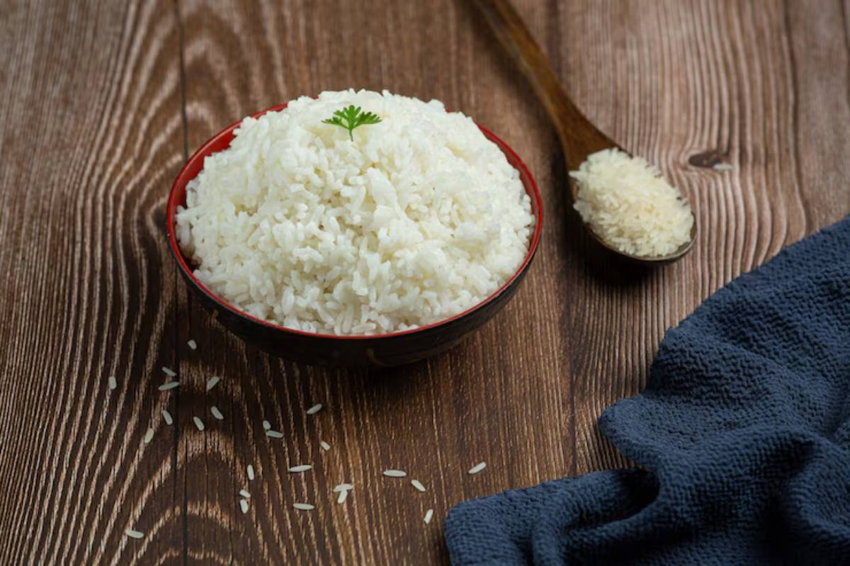 A bowl of fluffy white rice garnished with a sprig of cilantro, accompanied by a wooden spoon and a cloth napkin on a wooden surface.