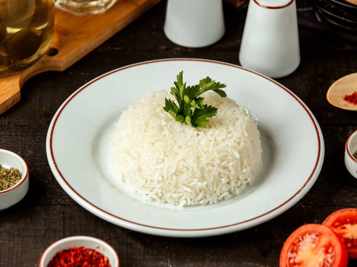 A plate of fluffy, white rice garnished with a sprig of parsley, surrounded by spices and fresh tomatoes on a rustic wooden table.