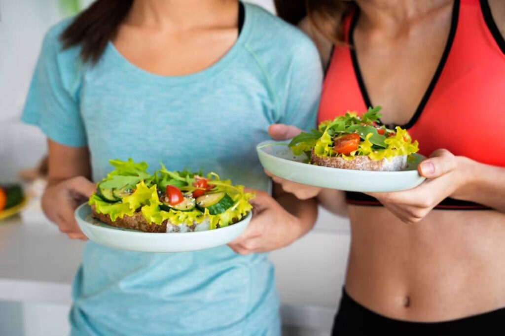 Two women in athletic wear hold plates of healthy vegetable-topped bread with lettuce, tomatoes, and cucumbers, smiling in a bright kitchen.