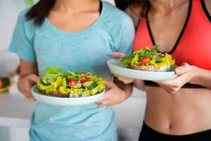 Two women in athletic wear hold plates of healthy vegetable-topped bread with lettuce, tomatoes, and cucumbers, smiling in a bright kitchen.