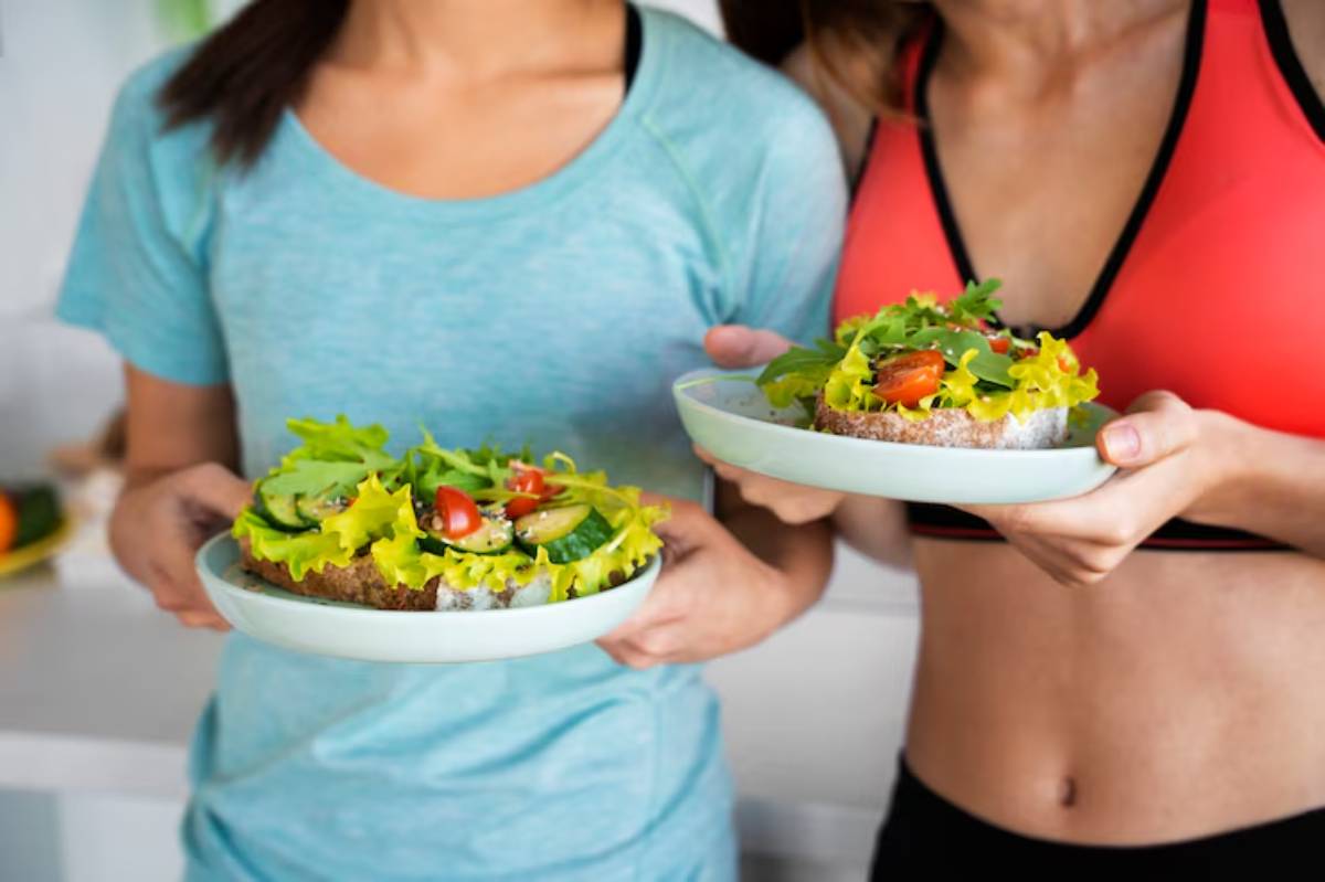 Two women in athletic wear hold plates of healthy vegetable-topped bread with lettuce, tomatoes, and cucumbers, smiling in a bright kitchen.