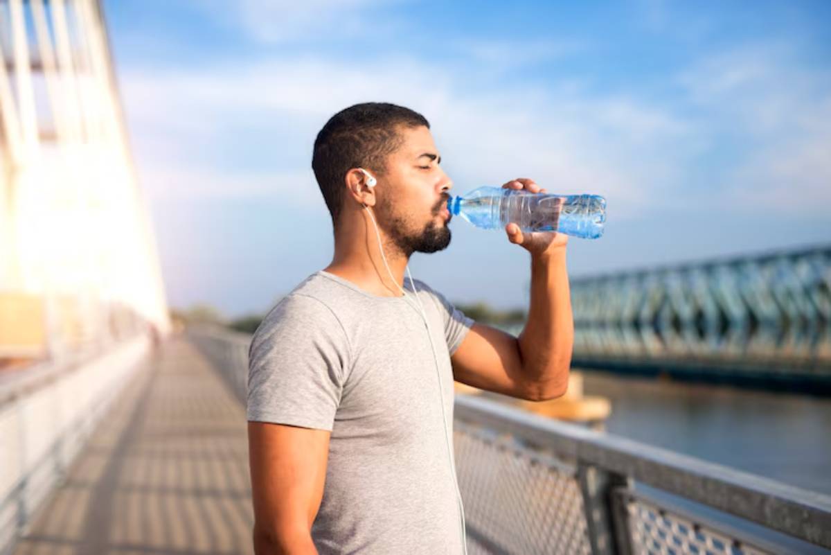 A person in a gray t-shirt drinks water from a bottle while standing on a bridge, with a scenic waterfront view.