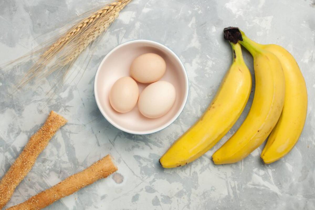 A bowl with three eggs, three bananas, and sesame sticks, surrounded by a stalk of wheat, on a gray surface.