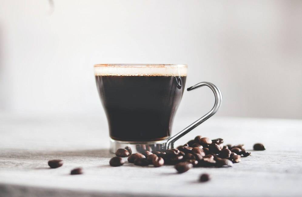 A sleek glass coffee cup filled with dark brew, with scattered coffee beans on a light wooden surface.