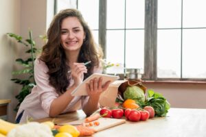 A woman with curly hair writes on a tablet while surrounded by fresh vegetables and fruits on a wooden kitchen table.