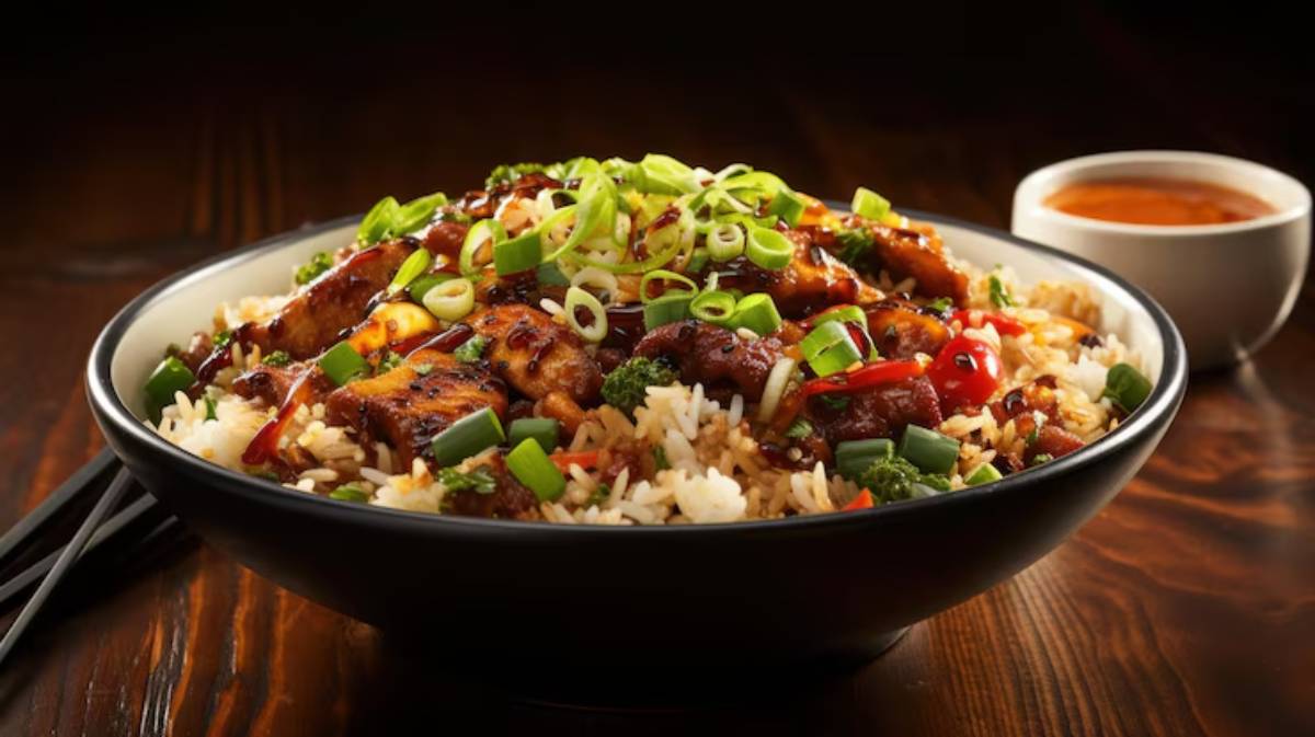 A vibrant bowl of fried rice topped with green onions, grilled chicken, and colorful vegetables, alongside a small cup of sauce.