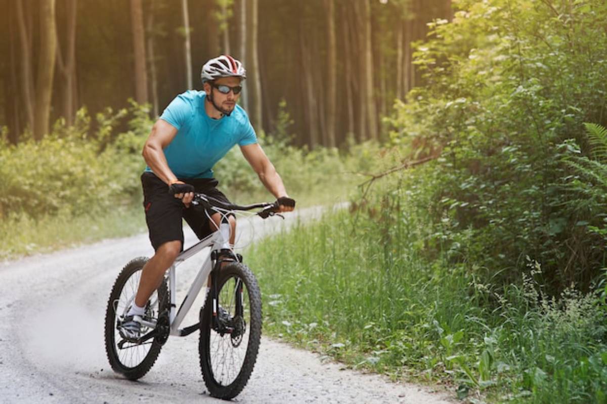 A cyclist in a blue shirt rides on a gravel path surrounded by lush greenery and trees, kicking up dust behind him.