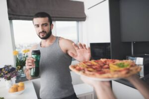 A man in a gray tank top gestures to decline a pizza while holding a green smoothie in a modern kitchen filled with flowers.
