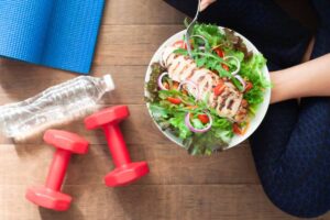 A person sitting on the floor with a bowl of grilled chicken salad topped with fresh vegetables, surrounded by a yoga mat, a water bottle, and red dumbbells.