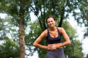 A woman in athletic wear stands outdoors, holding her stomach, with trees and soft natural light in the background.
