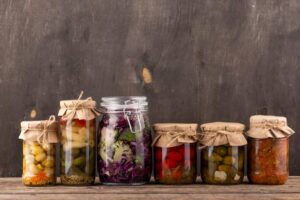 A collection of six jars with pickled vegetables and preserved foods, arranged on a wooden table against a rustic backdrop.