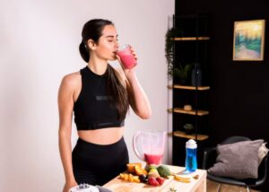A woman in athletic wear drinks a pink smoothie, surrounded by a blender and colorful fruits on a wooden table in a cozy kitchen setting.