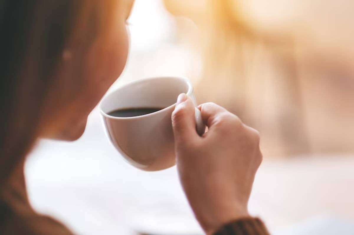 A woman holds a small white cup of coffee, gazing thoughtfully with a warm, soft background.