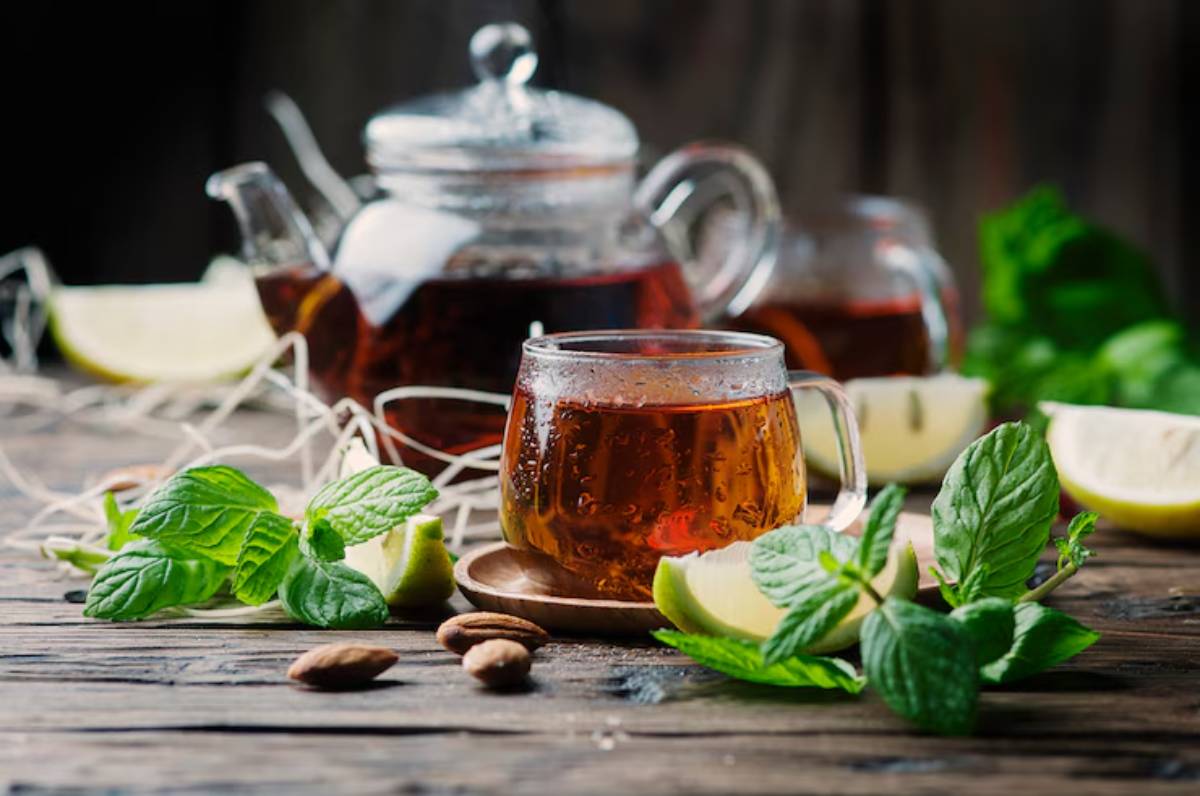 A glass of iced tea sits on a wooden table, surrounded by mint leaves, lemon slices, and almonds, with a teapot in the background.