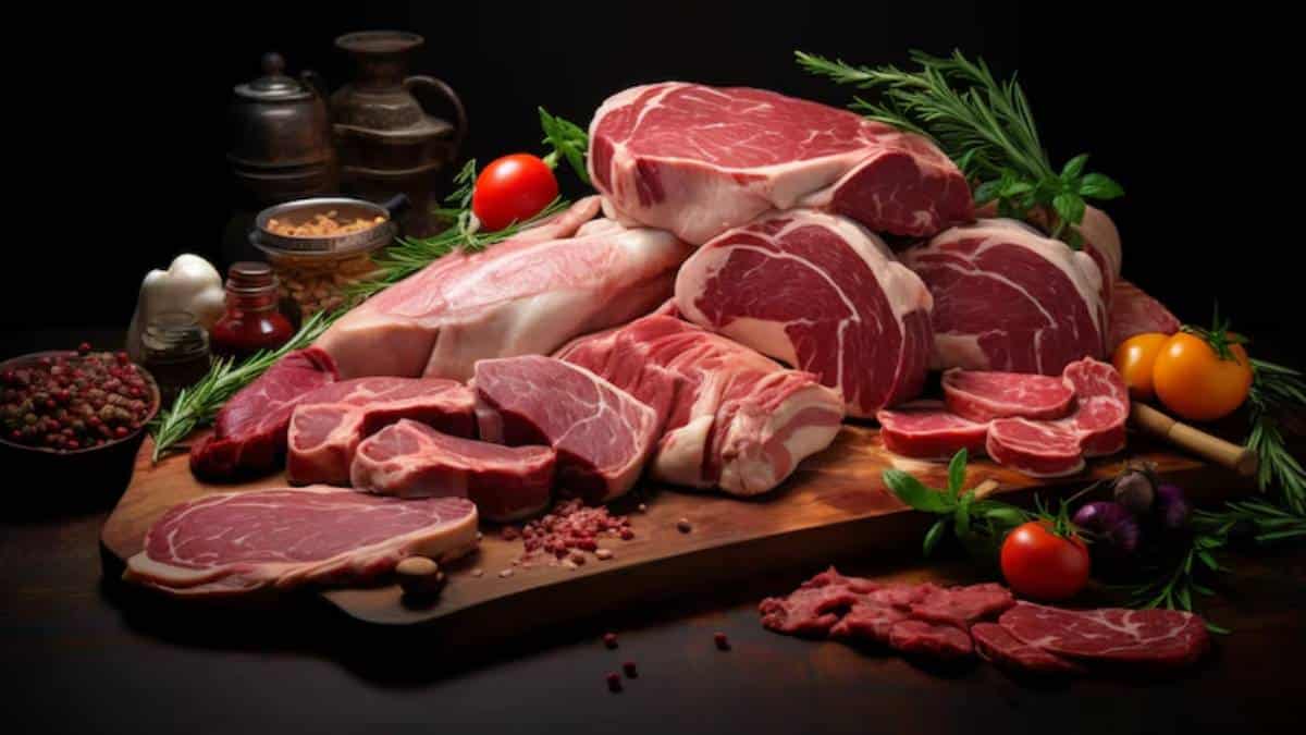 A variety of fresh, raw meats arranged on a wooden board, surrounded by herbs, spices, and colorful vegetables in a dark setting.