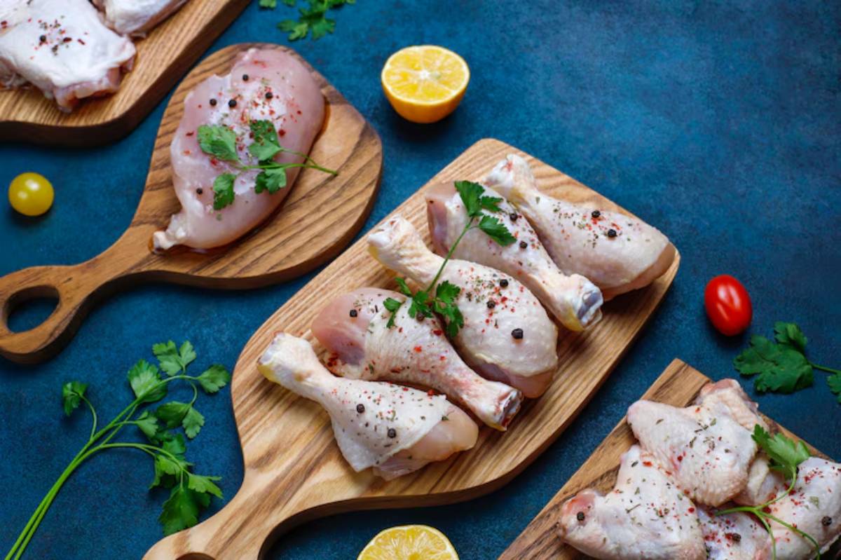 Fresh chicken pieces seasoned with herbs, arranged on wooden platters, alongside lemon and cherry tomatoes on a blue background.