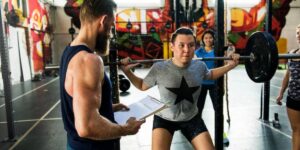 Two people are exercising in a bright indoor gym; one is in a wheelchair lifting weights with support from the other.
