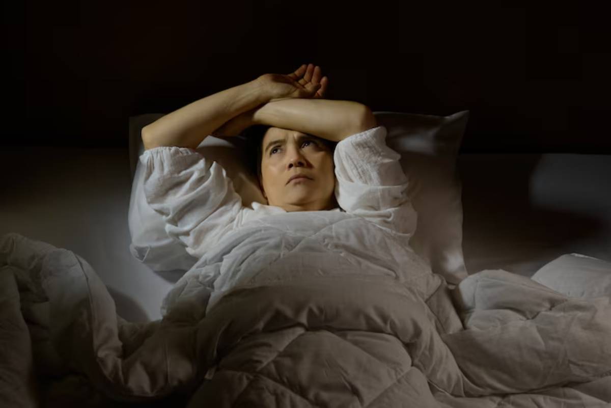 A person lies in bed, arms stretched above their head, partially covered by a white quilt in a dimly lit room.
