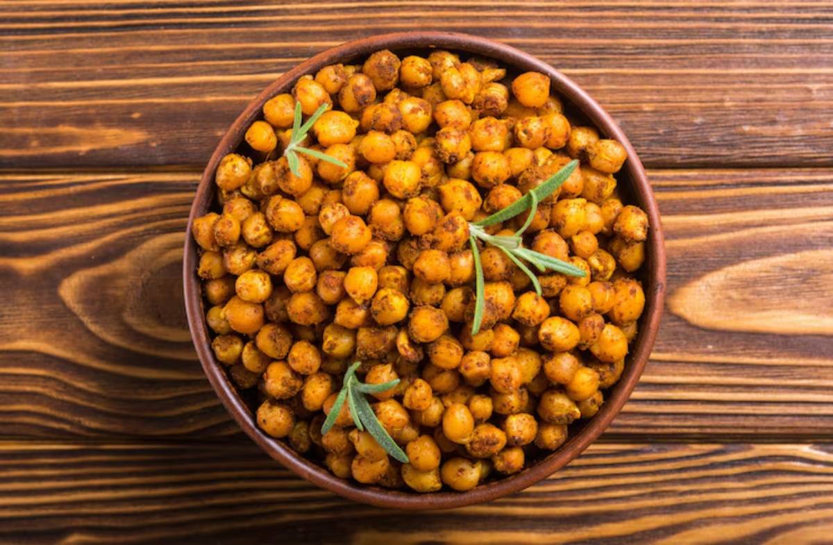 A brown bowl filled with seasoned, golden roasted chickpeas topped with fresh green herbs on a wooden surface.