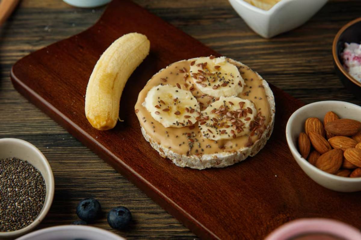 A wooden plate features a rice cake topped with peanut butter, banana slices, chia seeds, and flaxseeds, alongside a banana and almonds.