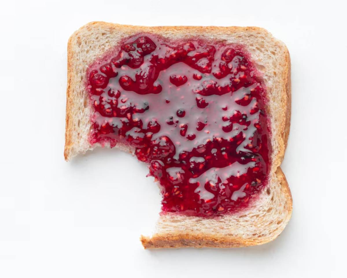 A slice of whole wheat bread topped with bright red raspberry jam, featuring a bite taken out from the lower left corner.