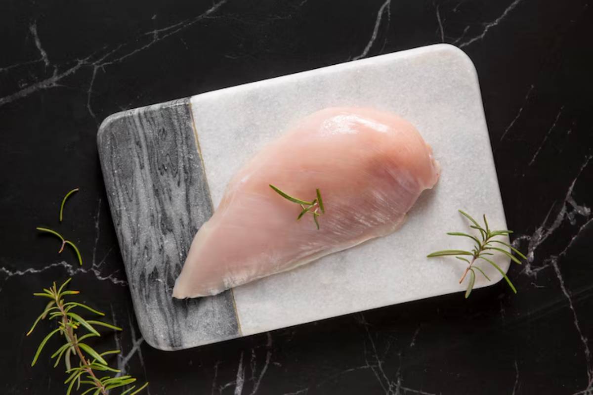 Raw chicken breast on a marble cutting board, garnished with fresh rosemary, against a dark background.