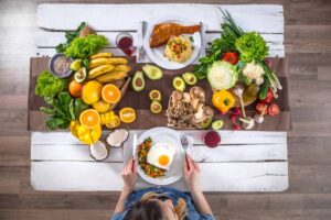 A woman prepares to eat a vibrant meal of veggies, pasta, and fish, surrounded by fresh fruits and vegetables on a rustic table.