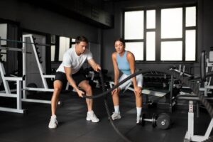 A person sits on the gym floor, drinking water from a bottle, with a barbell and weights nearby. The gym is bright and spacious.