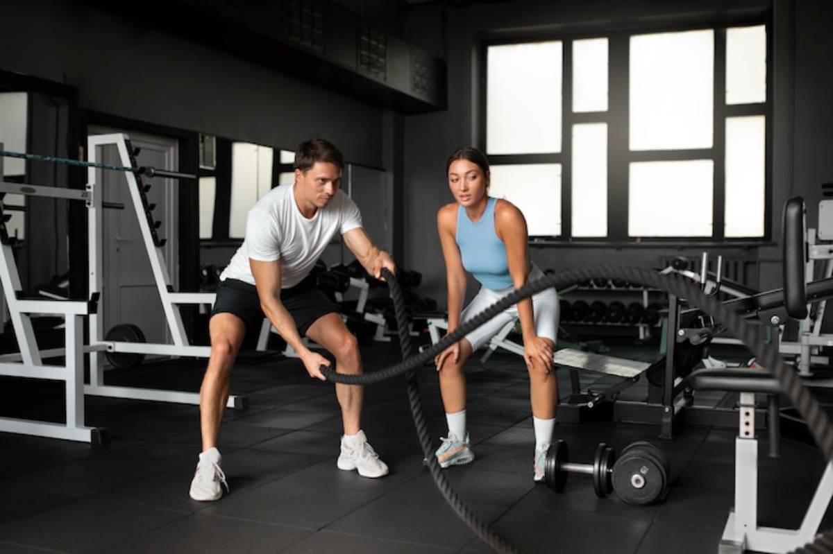 A person sits on the gym floor, drinking water from a bottle, with a barbell and weights nearby. The gym is bright and spacious.